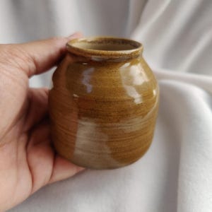 May include: A small, brown ceramic vase with a rounded shape and a smooth, glossy finish. The vase has a subtle, light brown glaze that gives it a warm, earthy tone.