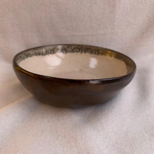 May include: A white ceramic bowl with a brown rim and a green speckled glaze. The bowl is sitting on a white surface.
