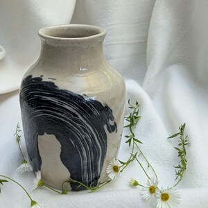 May include: A ceramic vase with a beige base and a bold, swirling black design. The vase has a rounded body and a narrow neck. Delicate white flowers with yellow centres and green stems are arranged around the base of the vase.