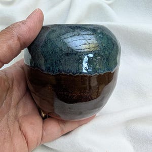 May include: A handmade ceramic bowl with a rounded shape. The top half is a dark teal color, while the bottom half is a rich brown. The bowl has a glossy finish and appears to be held in a hand.