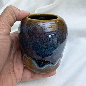 May include: A small, handcrafted ceramic vase with a glossy finish. The vase features a blend of brown, blue, and teal hues, creating a unique, marbled effect. The opening is circular, and the base has a wavy, textured design.
