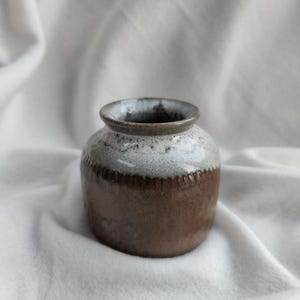 May include: A small, brown ceramic vase with a white glaze on the top half. The vase has a rounded body and a slightly flared lip.