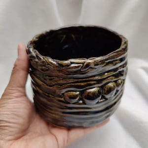 May include: A handmade ceramic pot with a dark, glossy interior and a textured exterior. The exterior has horizontal ridges and a series of rounded, raised accents. The colour is a blend of brown and bronze, with a shiny finish. The pot is held in a hand.