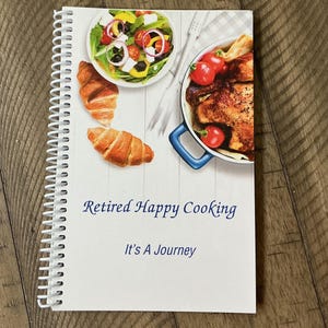 Op de afbeelding: Een spiraalgebonden kookboek met de titel "Retired Happy Cooking" met de slogan "It's A Journey". De omslag toont afbeeldingen van voedsel, waaronder een salade, croissants en een gerecht met gebraden kip en tomaten. Het boek ligt op een houten oppervlak.