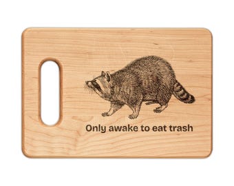 May include: A rectangular light brown wooden cutting board with a rounded handle cutout on the left side. The board features a black engraved illustration of a raccoon and the text "Only awake to eat trash" centered below the animal.