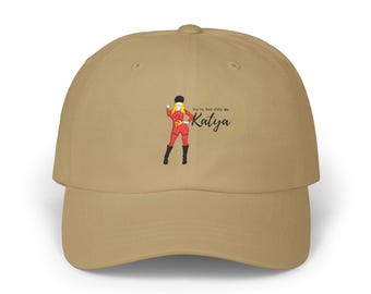 May include: A tan baseball cap featuring a graphic of a blonde figure in a red outfit, black hat, and black boots. The text reads "You're Best Calls Me Katya" in black script. The cap has a structured crown, a curved brim, and a button on top.