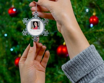 May include: A silver-toned snowflake-shaped Christmas ornament featuring a circular centre insert. The insert displays a cartoon illustration of a woman with dark hair and glasses wearing a pink top, with the text DO NOT FEED! printed below.