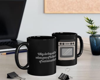 May include: Two black ceramic mugs. One mug features a graphic of a grey oven and the text, "Why do they call it oven when you of in the cold food of out hot eat the food". The mugs have a standard handle and a cylindrical shape.