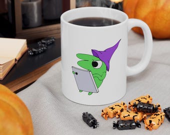 May include: A white ceramic mug featuring a graphic of a green creature wearing a purple pointed hat and holding a grey rectangular tablet. The mug has a standard handle and is filled with a dark liquid, showcasing a clean and simple design.