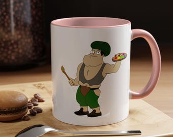 May include: A white ceramic mug with a light pink interior and handle, featuring a cartoon illustration of a muscular character wearing a green cap, dark grey tank top, and green shorts, holding a paintbrush and a colorful artist palette.