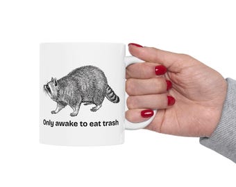 May include: A white ceramic mug featuring a black and white line art illustration of a raccoon walking. Below the raccoon, the text reads, "Only awake to eat trash" in a simple black sans-serif font. The mug has a standard handle on the right side.