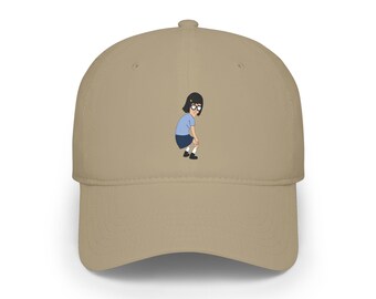 May include: A tan baseball cap featuring a small, centered graphic of a cartoon character with black hair, glasses, a light blue shirt, and a dark blue skirt, depicted in a hunched posture with hands resting on knees.