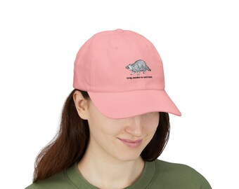 May include: A light pink baseball cap featuring a black and white line art illustration of a raccoon. Below the raccoon, the text reads "Only awake to eat trash" in a simple black sans-serif font. The cap has a curved brim and a structured crown.