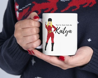 May include: A white ceramic mug featuring a graphic illustration of a blonde figure wearing a red outfit, black boots, and a black hat. The figure has a gold hammer and sickle emblem on the chest. The text reads, "You're Dad Calls Me Katya."