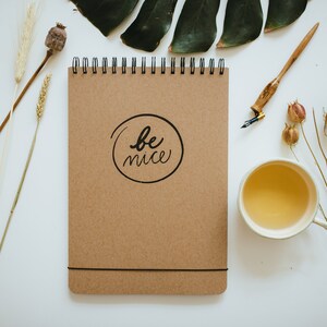 May include: A brown spiral-bound notebook with the words "be nice" in a black circle. The notebook is on a white surface, along with a cup of tea, a pen, and dried plants.