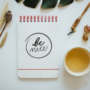 May include: A white notepad with the words "be nice" in a black circle, a cup of tea, a calligraphy pen, and dried flowers are arranged on a white surface. The notepad has a red spiral binding and a red elastic band.