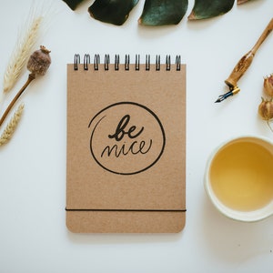 May include: A spiral-bound notepad with the words "be nice" in a black circle. The notepad is brown and sits on a white surface, alongside a cup of tea, a pen, and dried plants.