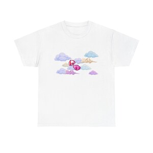 May include: A white short-sleeve t-shirt featuring a central graphic of a small pink character floating with a red and white striped umbrella amidst a cluster of soft, pastel-colored clouds in shades of light blue, lavender, and cream.