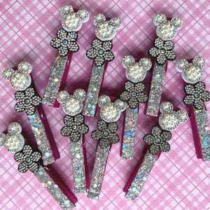 May include: A collection of decorative hair clips with a Mickey Mouse head design. Each clip features a pearl-like head, a flower-shaped embellishment, and a glittery rectangular base. The clips are arranged on a pink checkered surface.