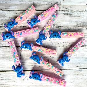 May include: A collection of light pink wooden clothespins decorated with colorful sprinkles and a small blue character figurine attached to each one. The character features large ears, a wide smile, and a round nose, consistent across all pieces.