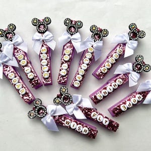 May include: A collection of decorative purple glitter pens with white bows and rhinestone Mickey Mouse-shaped accents. Each pen has a different positive word spelled out in white letters, such as "SOAR", "SHINE", and "DREAM".