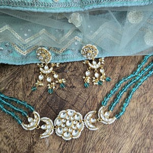 Op de afbeelding: Een turquoise kralenketting met een gouden hanger met een grote, ronde, witte steen omringd door kleinere stenen. De hanger heeft ook twee halvemaanvormen aan weerszijden. De ketting wordt geleverd met bijpassende oorbellen met een vergelijkbaar ontwerp.