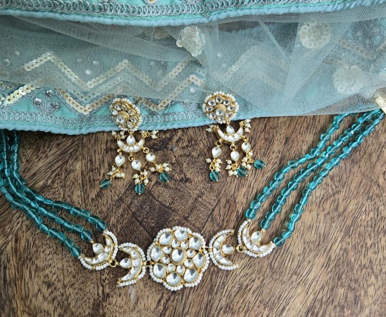 Op de afbeelding: Een turquoise kralenketting met een gouden hanger met een grote, ronde, witte steen omringd door kleinere stenen. De hanger heeft ook twee halvemaanvormen aan weerszijden. De ketting wordt geleverd met bijpassende oorbellen met een vergelijkbaar ontwerp.