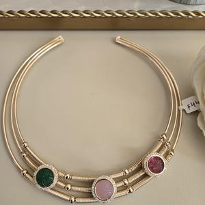 May include: A gold-toned necklace with three circular bands. The necklace features three gemstone pendants: green, pink, and red, each surrounded by small, clear stones. The gemstones have carved floral designs. The necklace is displayed on a neutral surface.