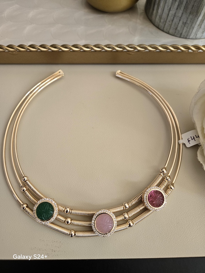May include: A gold-toned necklace with three circular bands. The necklace features three gemstone pendants: green, pink, and red, each surrounded by small, clear stones. The gemstones have carved floral designs. The necklace is displayed on a neutral surface.