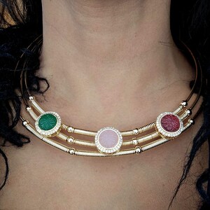 May include: A gold-toned necklace with three circular gemstone pendants. The pendants are green, pink, and red, each surrounded by a ring of small, clear crystals. The necklace has a multi-strand design.