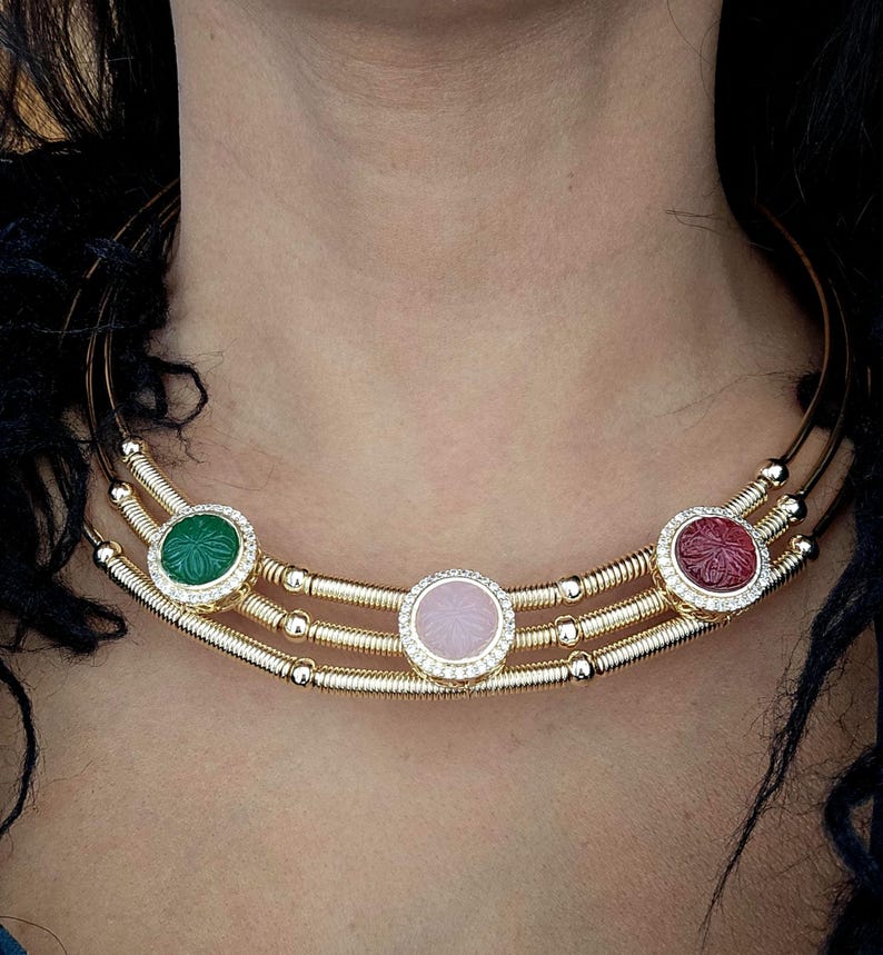 May include: A gold-toned necklace with three circular gemstone pendants. The pendants are green, pink, and red, each surrounded by a ring of small, clear crystals. The necklace has a multi-strand design.