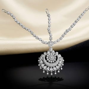 May include: A silver-toned headpiece with a floral and crescent moon design, adorned with numerous clear gemstones. The headpiece features delicate chains with small, sparkling accents, and a central pendant with dangling teardrop-shaped gems.