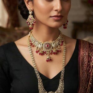 Peut inclure: Un ensemble de bijoux doré composé d'un collier ras-du-cou et d'un long collier pendentif, ornés de filigrane complexe, de pierres blanches incrustées et d'accents rouge foncé. Des boucles d'oreilles assorties complètent l'ensemble avec des perles.