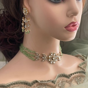 Op de afbeelding: Een gouden chokerketting met een groot, rond, wit stenen middelpunt en twee halve maan accenten. De ketting is gemaakt van kleine, groene kralen en wordt gedragen door een mannequin.