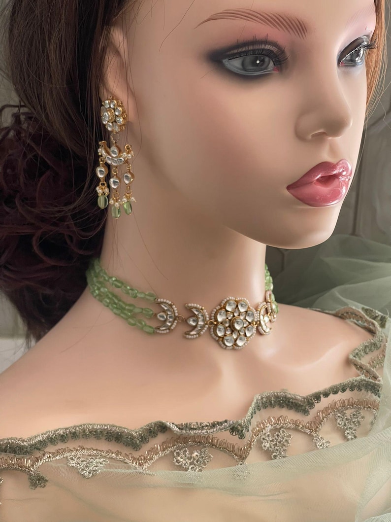 Op de afbeelding: Een gouden chokerketting met een groot, rond, wit stenen middelpunt en twee halve maan accenten. De ketting is gemaakt van kleine, groene kralen en wordt gedragen door een mannequin.