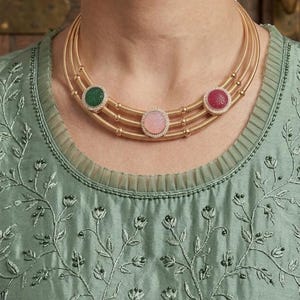 May include: A gold necklace with three strands, each adorned with a gemstone pendant. The pendants feature a green, pink, and red stone, each surrounded by small, clear stones. The necklace is worn over a green embroidered top.