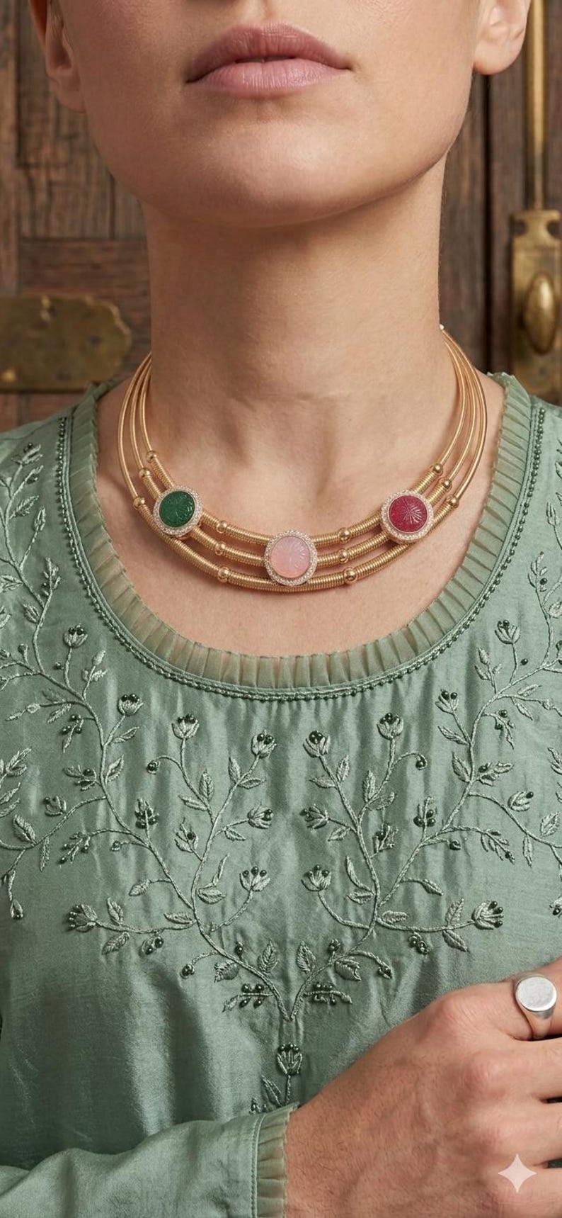 May include: A gold necklace with three strands, each adorned with a gemstone pendant. The pendants feature a green, pink, and red stone, each surrounded by small, clear stones. The necklace is worn over a green embroidered top.