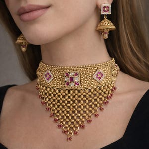 May include: Gold-toned necklace and earring set featuring intricate detailing and ruby-colored accents. The choker-style necklace has a square pendant with a central ruby and diamond-like embellishments. The earrings have a similar design.