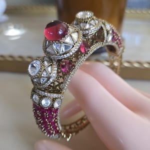 May include: A gold-toned hinged bangle bracelet featuring an intricate openwork metal design. The piece is adorned with multiple faceted clear stones, round cabochon-style red stones, and small pavé-set crystals throughout the surface.