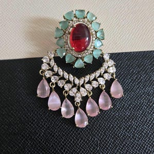 May include: An ornate earring featuring a large oval red gemstone surrounded by teal-colored stones. Below, a crescent of clear and white stones supports dangling pink teardrop-shaped gems. The earring has a gold-colored metal setting.