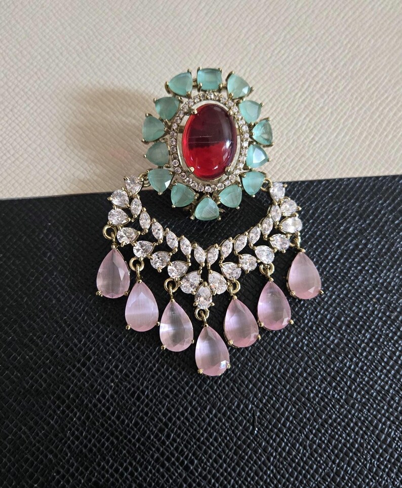 May include: An ornate earring featuring a large oval red gemstone surrounded by teal-colored stones. Below, a crescent of clear and white stones supports dangling pink teardrop-shaped gems. The earring has a gold-colored metal setting.