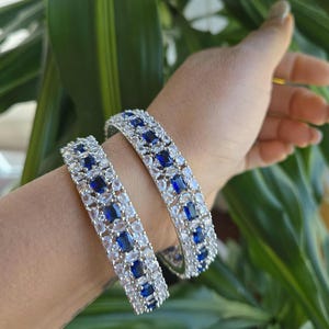 May include: Two silver bangles with blue and white stones. The bangles are stacked on a person's wrist.