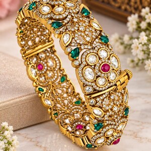 May include: A gold hinged bangle bracelet featuring an intricate filigree design. The surface is embellished with clear white crystals, teardrop-shaped green stones, and round pink stones arranged in a floral pattern across the entire band.