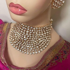 May include: A gold-coloured choker necklace and matching earrings, both set with numerous clear, sparkling stones. The jewellery is displayed on a mannequin with pink lips and a pink and gold garment.