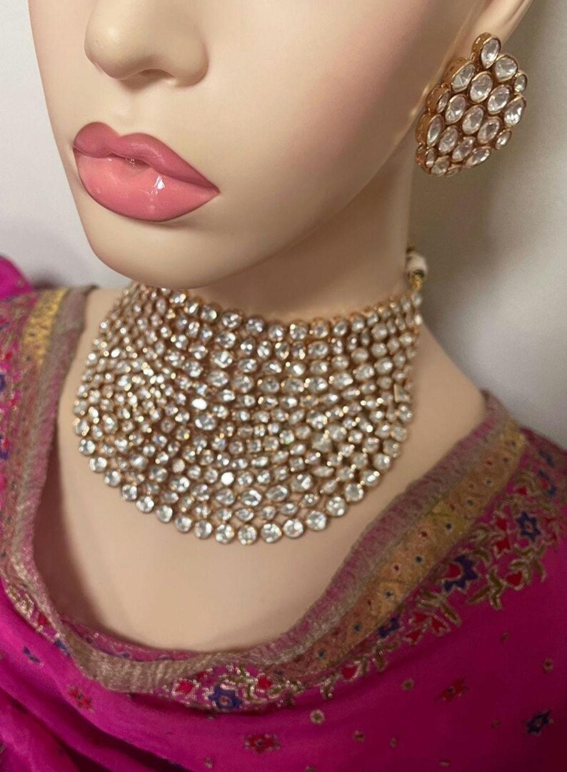 May include: A gold-coloured choker necklace and matching earrings, both set with numerous clear, sparkling stones. The jewellery is displayed on a mannequin with pink lips and a pink and gold garment.
