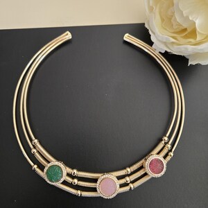 May include: A gold-toned collar necklace with three circular gemstone accents. The necklace features three parallel bands, with the gemstones in green, pink, and red. Each gemstone is surrounded by a halo of small, clear crystals. The necklace is displayed on a black surface.