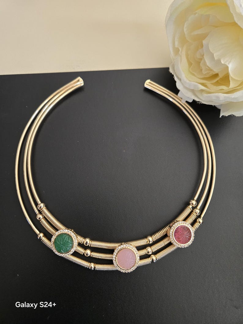 May include: A gold-toned collar necklace with three circular gemstone accents. The necklace features three parallel bands, with the gemstones in green, pink, and red. Each gemstone is surrounded by a halo of small, clear crystals. The necklace is displayed on a black surface.