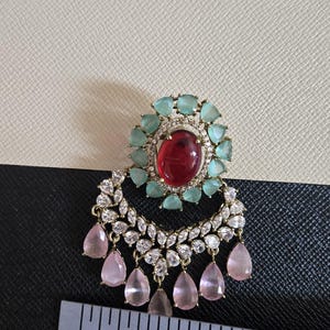 May include: An ornate earring with a large red oval stone at the center, surrounded by light blue stones. Below, a crescent shape is adorned with clear stones and pink teardrop-shaped gems. A ruler is placed below the earring.
