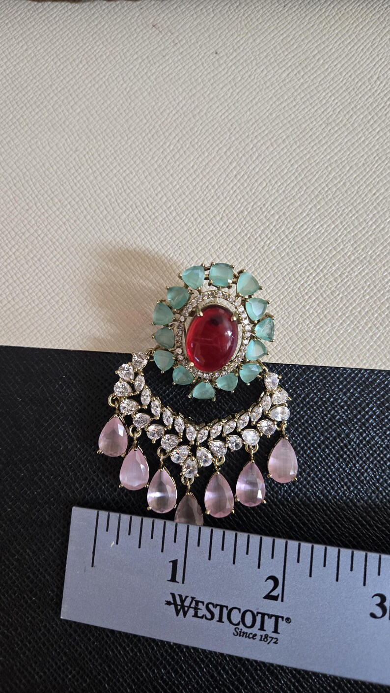 May include: An ornate earring with a large red oval stone at the center, surrounded by light blue stones. Below, a crescent shape is adorned with clear stones and pink teardrop-shaped gems. A ruler is placed below the earring.