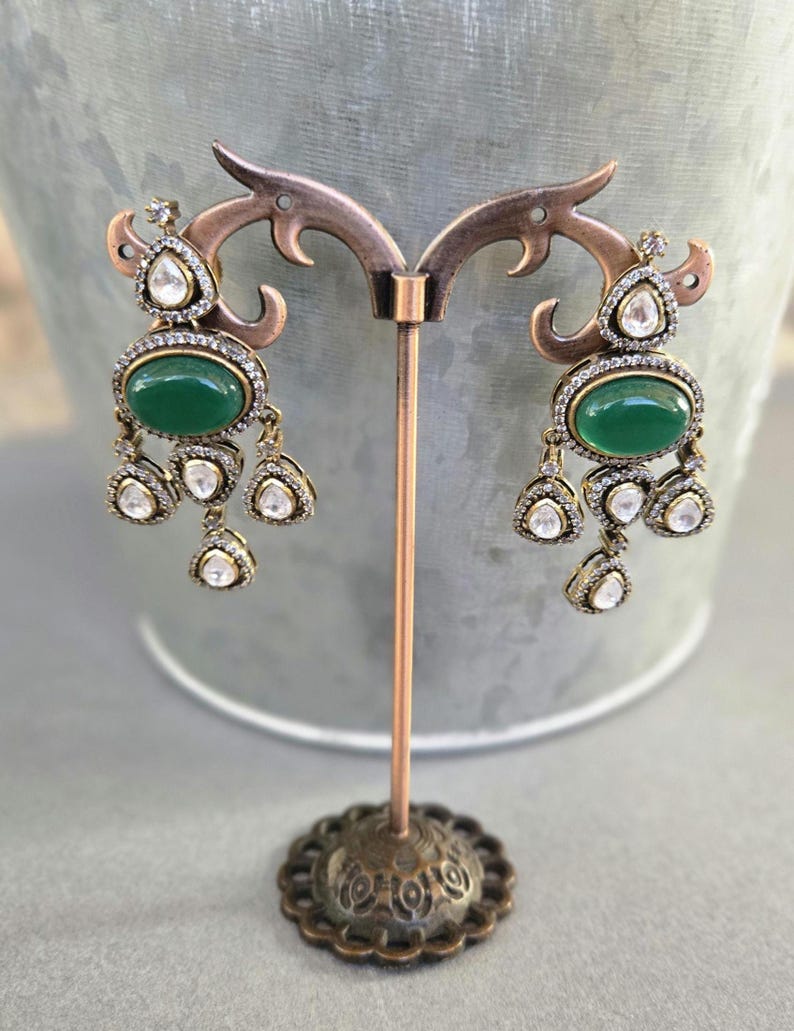 Puede incluir: Un par de pendientes colgantes con una piedra ovalada verde y brillantes transparentes. Los pendientes est&aacute;n en un soporte de metal.